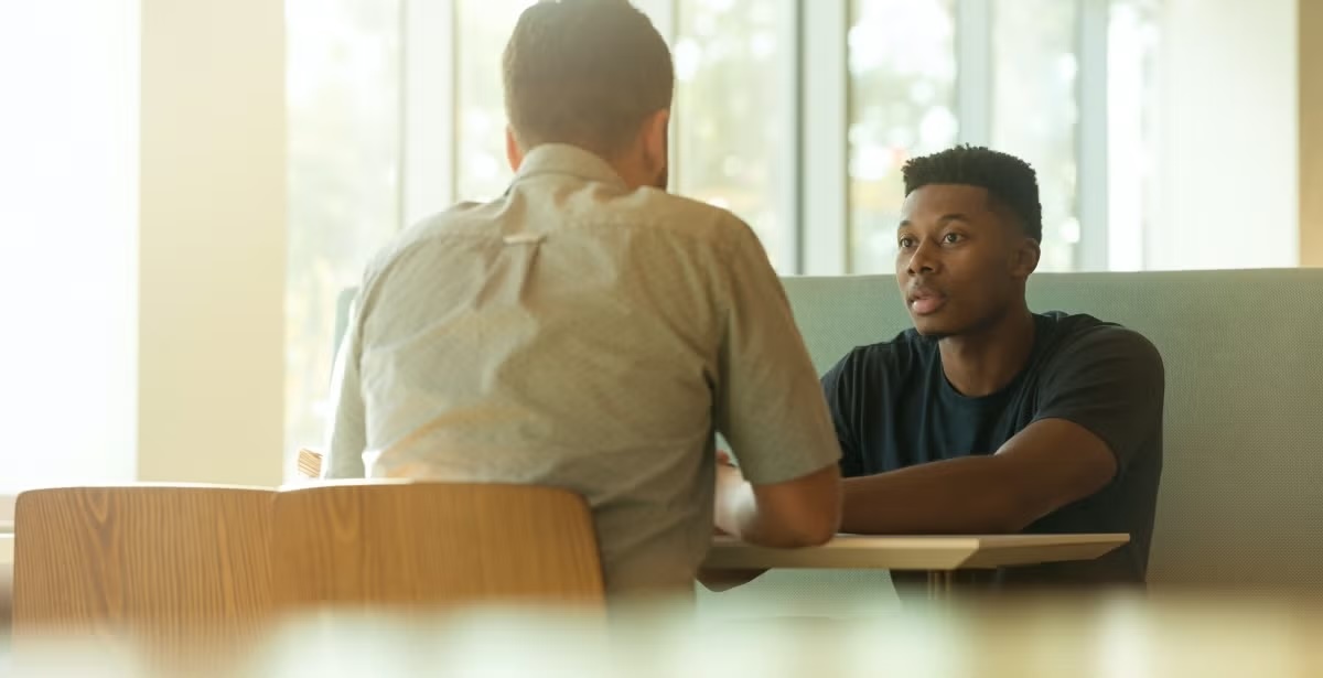 Two students sitting at a table in a café, having a conversation by large windows with natural light.