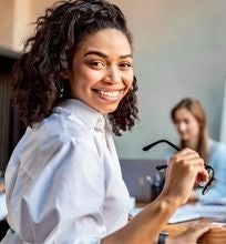 A smiling woman holding glasses during a business meeting with colleagues working in the background.