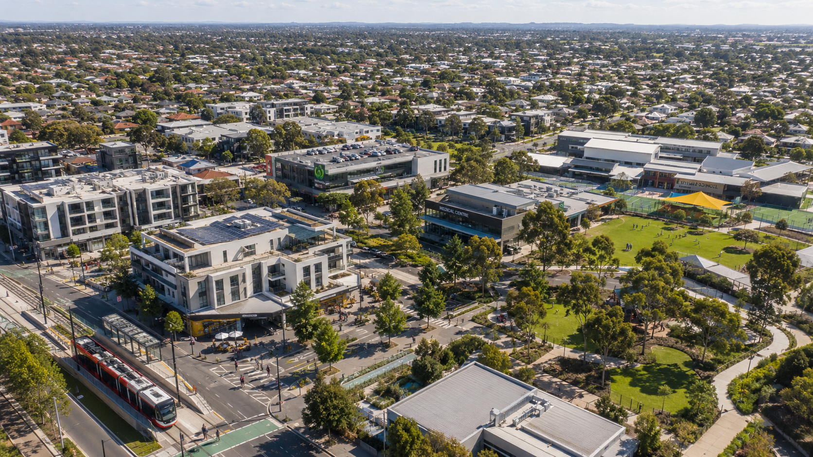 Aerial view of a compact Australian neighbourhood with medium-density housing, light rail, local shops, a medical centre, school, parks and surrounding suburban sprawl.
