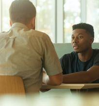 Two students sitting at a table in a café, having a conversation by large windows with natural light.