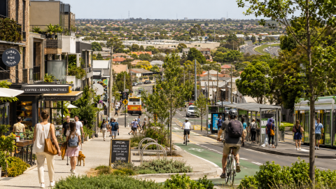 A walkable Australian neighbourhood with pedestrians, cyclists, public transport, local shops, trees and medium-density housing, contrasting with suburban sprawl in the distance.