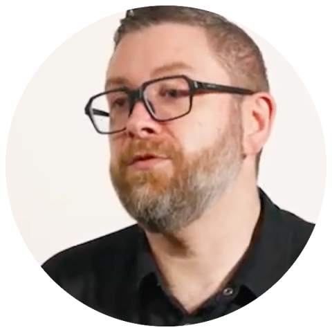 A man with glasses and a beard wearing a black shirt is looking directly at the camera.