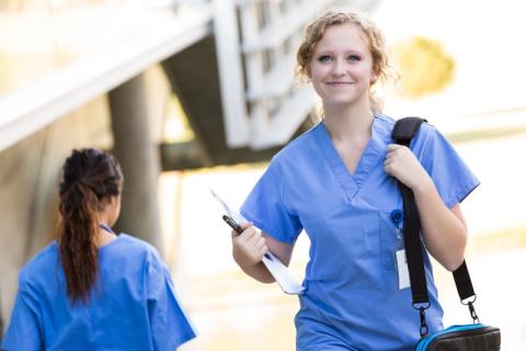 A Master of Advanced Nursing graduate walking outside a hospital carrying a bag and a clip board.