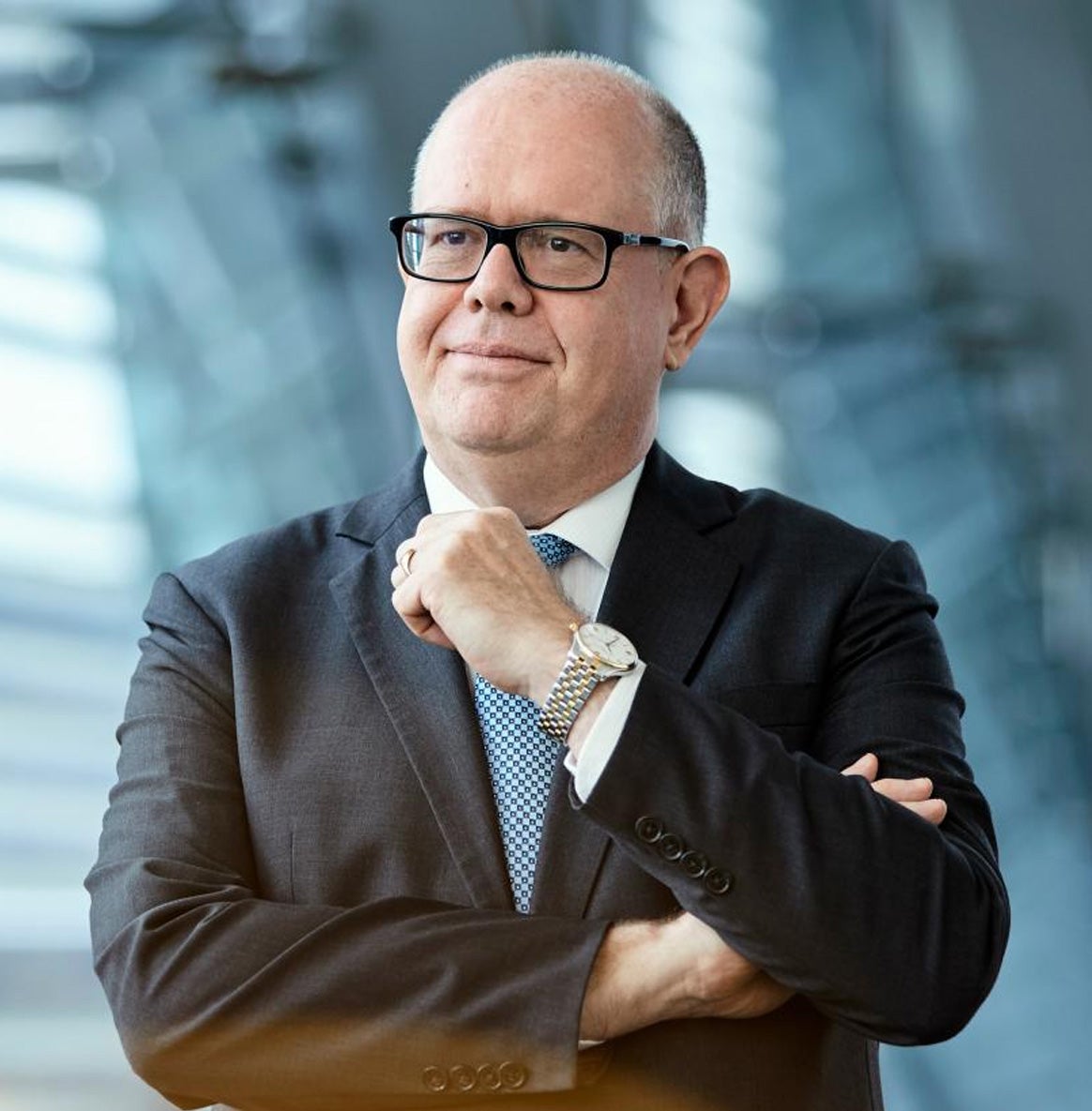 A man wearing glasses and a suit stands confidently in front of a large window, showcasing a professional demeanor.