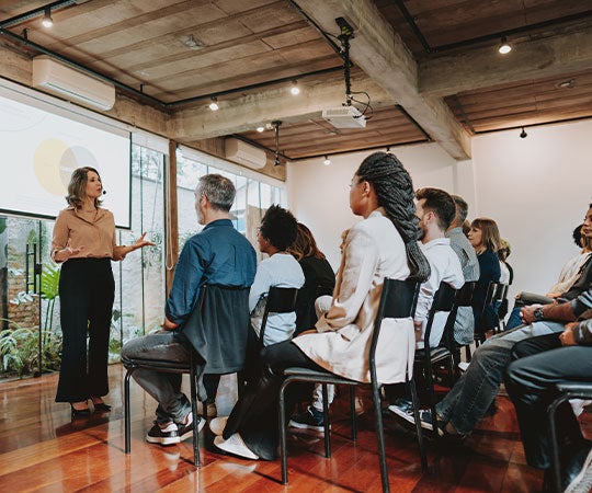 1. A woman stands at a podium, presenting to an audience seated in rows, engaged and attentive.