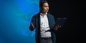 A man stands in front of a screen, holding a laptop, engaged in a presentation or discussion.