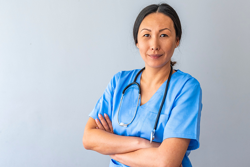 A woman in a blue scrub suit stands with her arms crossed, displaying a confident and professional demeanor.