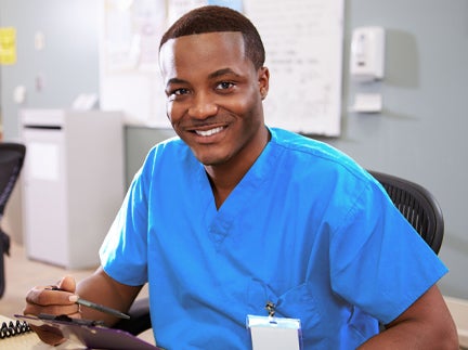 A smiling male nurse in scrubs sits at a desk, conveying a friendly and approachable demeanor in a healthcare setting.