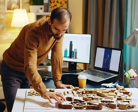 A man working on an architectural model with urban design plans displayed on computer screens in a home office.