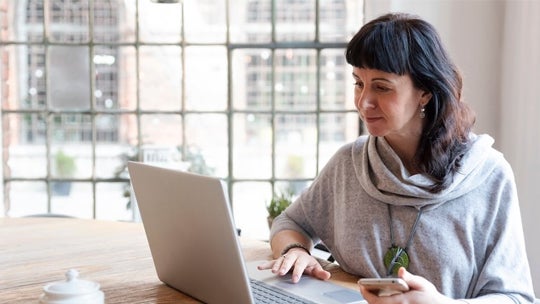 A woman seated at a table, focused on her laptop, with a calm expression in a well-lit room.