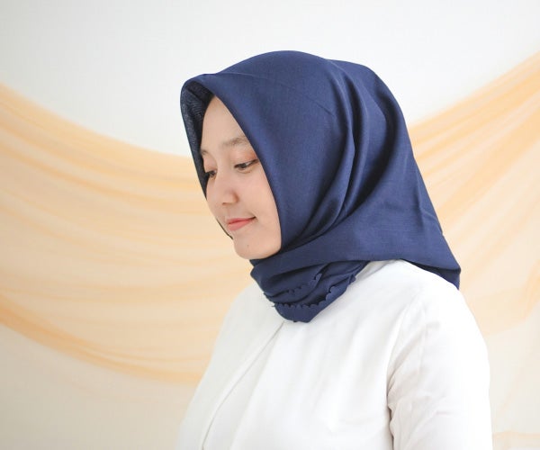 A woman wearing a blue hijab, smiling softly, with her hair partially visible beneath the fabric.
