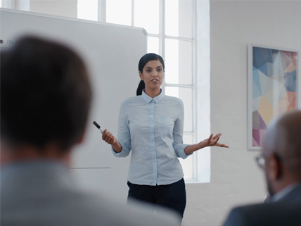 A professional woman giving a presentation in a modern conference room with a whiteboard.