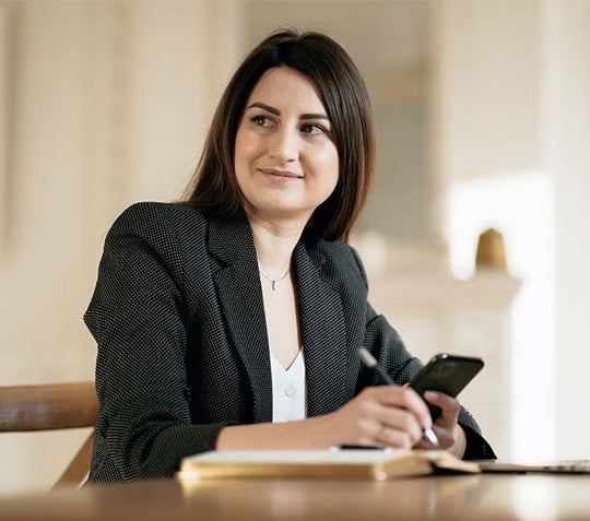 A woman in a business suit sits at a desk, focused on her work with a laptop and documents in front of her.