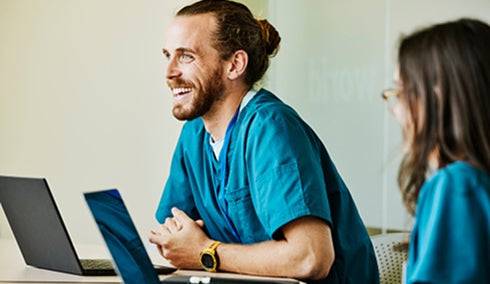 A man and woman in scrubs sit at a table, working on laptops and engaged in discussion.