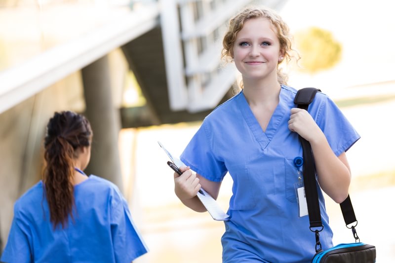 A Master of Advanced Nursing graduate walking outside a hospital carrying a bag and a clip board.