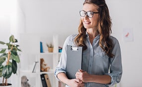 A woman wearing glasses holds a clipboard while standing in an office environment.