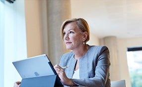 A woman in a business suit sits at a table, focused on her laptop, in a professional setting.