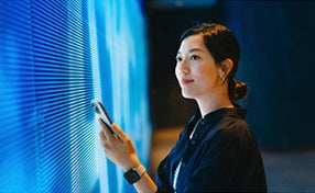 A woman interacts with a smartphone while standing in front of a large digital screen displaying vibrant graphics.