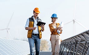 Two individuals in hard hats walking alongside a row of solar panels under a clear blue sky.