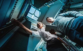Two individuals standing in a server room, surrounded by rows of servers and networking equipment.
