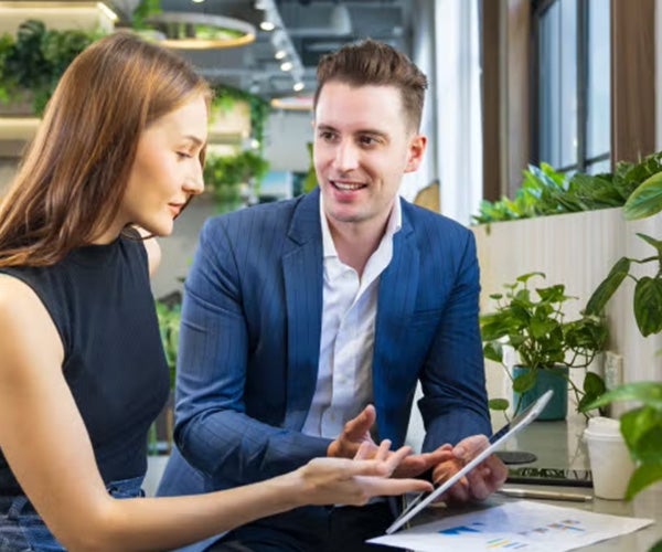 Two colleagues having a discussion while seated next to green plants in a bright, modern office setting.