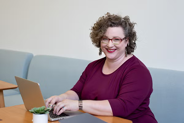 A smiling woman with a curly hair working in front a laptop in a cafe.