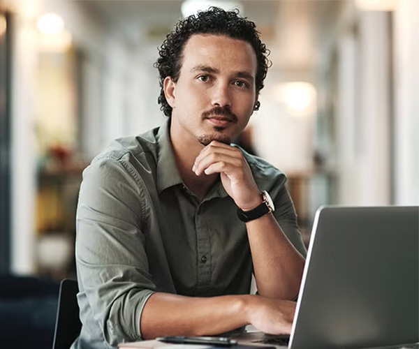 Man in a green shirt sitting at a desk, resting his chin on his hand while looking thoughtfully at the camera with a laptop in front of him.