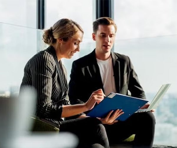 Two colleagues having a discussion in an office space, one holding a blue folder while seated.