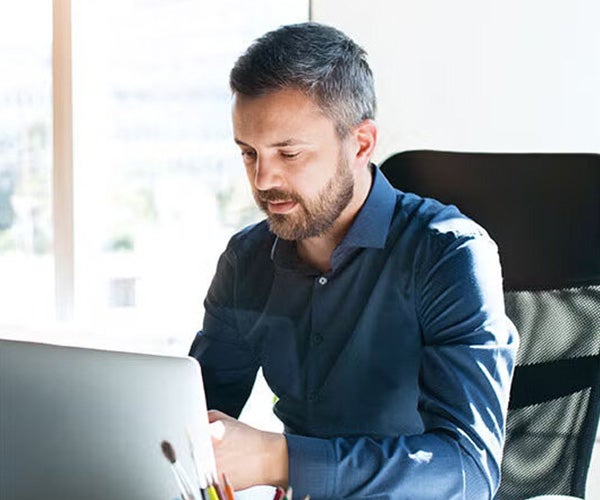 A professional man with short gray hair and a beard works on a laptop in a bright, modern office.