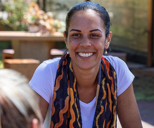 1. A woman smiles while seated outdoors among a group of people, enjoying a social gathering in a pleasant environment.