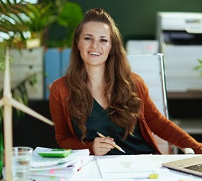 A woman seated at a desk, working on a laptop surrounded by papers.